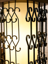 Load image into Gallery viewer, Close-up of a decorative metal lantern or screen with vertical black scrollwork bars in front of a glowing warm white cylindrical light source.
