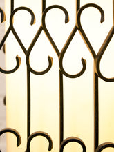 Load image into Gallery viewer, Close-up of a decorative wrought iron gate or window screen with repeating scrollwork and vertical bars, shown against a warm glowing background.

