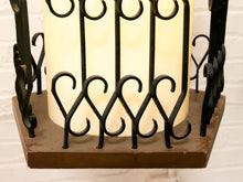 Load image into Gallery viewer, A close-up of a decorative lantern or light fixture with a glowing warm white cylindrical shade, black wrought-iron scrollwork, and a square brown wooden base against a white brick wall.
