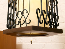 Load image into Gallery viewer, A close-up of a hanging lantern-style light fixture with a dark brown wooden frame, black wrought iron scrollwork, and a warm cream-colored glass shade. A pull chain hangs from the bottom center.
