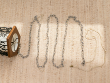 Load image into Gallery viewer, A silver metal link chain draped in a loose wave pattern on a beige woven fabric background, with a small portion attached to a decorative lantern on the left.
