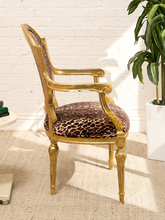 Load image into Gallery viewer, An elegant French-style armchair with a carved gold gilded frame and leopard-print upholstered seat and backrest, set against a white brick wall with a potted plant nearby.

