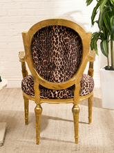 Load image into Gallery viewer, A vintage-style gold armchair with leopard-print upholstery, set against a white brick wall background with a potted plant on the right.
