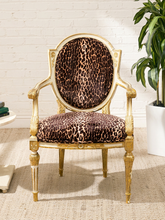 Load image into Gallery viewer, A classic French-style accent chair with a carved gold frame and leopard-print upholstery on the seat and oval backrest, set against a white brick wall background.
