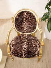Load image into Gallery viewer, An ornate gold-framed accent chair with leopard-print fabric upholstery, set on a beige woven rug against a white brick wall with a green plant in the background.
