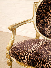 Load image into Gallery viewer, Close-up of an ornate baroque-style armchair with a gold gilded frame and leopard-print velvet upholstery, set against a white brick wall and light carpet.
