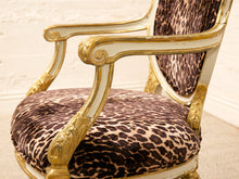 Load image into Gallery viewer, A close-up of an ornate armchair with a leopard-print upholstered seat and backrest, featuring carved gold-finished wooden arms and frame details against a light cream wall.
