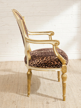 Load image into Gallery viewer, A vintage-style armchair with a gold-painted frame and leopard-print upholstered seat and backrest, set against a white brick wall and light beige rug.
