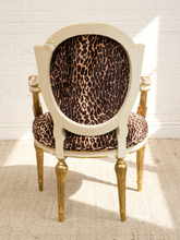 Load image into Gallery viewer, An elegant vintage-style armchair with a distressed cream frame, gold-finished legs, and leopard-print fabric upholstery, set against a white brick wall with soft sunlight and shadows.
