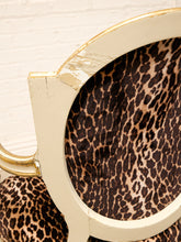 Load image into Gallery viewer, Close-up of a leopard-print upholstered chair with a distressed cream frame and gold trim, set against a white brick wall.
