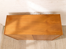 Load image into Gallery viewer, A mid-century modern wooden cabinet with a warm honey-toned wood grain finish, photographed from a top-down perspective against a white brick wall and textured rug.
