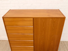 Load image into Gallery viewer, A mid-century modern wooden cabinet with five drawers on the left and a single wooden door on the right, finished in a warm honey-brown teak wood grain, set against a white brick wall.
