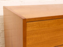 Load image into Gallery viewer, Close-up of a mid-century modern wooden chest of drawers with a warm honey-toned finish, showing the corner joint and drawer front against a white brick wall.
