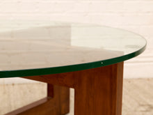 Load image into Gallery viewer, A close-up of a round glass-top table with a warm brown wooden base and a dark green beveled glass edge, set against a light brick wall background.
