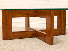 Load image into Gallery viewer, A mid-century modern wooden coffee table with a rich brown wood grain base and a clear glass top, set on a light beige textured rug against a white brick wall.

