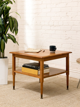 Load image into Gallery viewer, A mid-century modern wooden side table with a warm medium-brown finish, featuring a top surface and a lower shelf. The table is styled in a room setting with a white brick wall background, a potted green plant on the left, and books and a mug on top.
