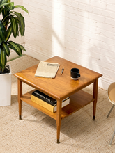 Load image into Gallery viewer, Vintage two-tier wood side table with square top, open lower shelf, tapered legs, warm wood grain, and brass-tone feet
