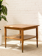 Load image into Gallery viewer, A mid-century modern wooden side table with a warm medium-brown finish, featuring a top surface, a lower shelf, and tapered legs with dark metal feet, set against a white brick wall and beige carpet.
