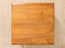 Load image into Gallery viewer, A square wooden board with rounded corners, showing a warm honey-brown wood grain pattern, placed on a textured beige woven mat.
