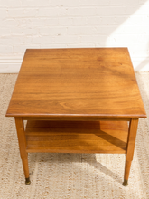 Load image into Gallery viewer, A mid-century modern wooden side table with a warm brown finish, featuring a square top, open lower shelf, and tapered legs, set on a woven rug against a white brick wall.
