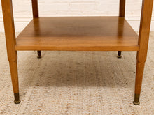 Load image into Gallery viewer, Close-up of the lower portion of a wooden two-tier side table or end table, showing the warm medium-brown wood grain of the shelves and legs, with brass-colored feet, set on a textured beige rug against a white brick wall.
