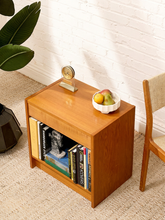 Load image into Gallery viewer, A small wooden side table or end shelf with a warm medium-brown finish, styled in a room corner with books, a small clock, a bowl of fruit, a white brick wall, and part of a chair visible.
