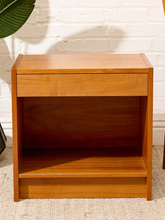 Load image into Gallery viewer, A mid-century modern wooden nightstand with a warm honey finish, featuring a single drawer and an open shelf, set against a white brick wall background.
