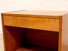 Load image into Gallery viewer, A close-up of a mid-century modern wooden side table or shelf with a single drawer, featuring a warm honey-toned wood finish and a white brick wall background.
