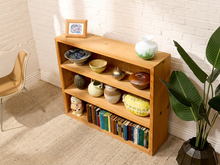 Load image into Gallery viewer, A light natural wood 4-tier bookshelf styled with books, ceramic vases, and a small picture frame, set against a white brick wall in a bright room.
