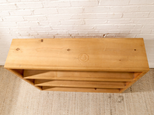 Load image into Gallery viewer, A light natural wood bookshelf with three open shelves, positioned against a white brick wall and a beige textured rug.
