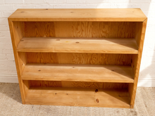 Load image into Gallery viewer, A three-tier wooden bookshelf with a natural light oak finish, standing against a white brick wall on a beige carpet.
