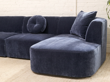 Load image into Gallery viewer, A modern modular sectional sofa upholstered in dark navy blue velvet, featuring a chaise lounge, a round tufted cushion, and a square back cushion, set against a white brick wall and beige carpet.
