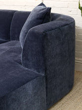 Load image into Gallery viewer, A close-up of a curved armchair upholstered in dark navy blue velvet fabric, featuring a matching throw pillow, set against a white brick wall and a woven rug.
