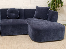 Load image into Gallery viewer, A modern sectional sofa in dark navy blue velvet, featuring a chaise lounge on the right, a round tufted back cushion, and a square throw pillow, set on a beige woven rug against a white brick wall with a green plant in the background.

