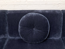 Load image into Gallery viewer, A round tufted throw pillow in dark navy blue velvet, resting on a matching dark navy velvet sofa in front of a white brick wall.
