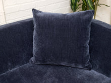 Load image into Gallery viewer, A square navy blue textured throw pillow resting on a matching navy blue upholstered sofa, with a white brick wall and a green plant in the background.
