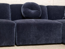 Load image into Gallery viewer, A close-up of a dark navy blue velvet sectional sofa with a matching round tufted throw pillow, set against a white brick wall background.
