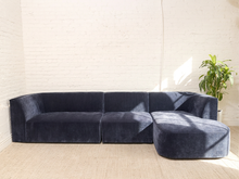 Load image into Gallery viewer, A dark navy blue velvet modular sectional sofa with a left-facing armrest and a right-facing ottoman, set against a white brick wall with a green potted plant on a light beige floor.
