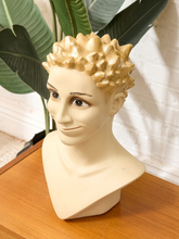 Load image into Gallery viewer, A decorative male bust sculpture with a glossy cream-colored torso and neck, a sculpted spiky blond hairpiece in a warm gold tone, and painted facial features, displayed on a wooden surface with a white brick wall and green leaves in the background.
