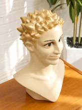 Load image into Gallery viewer, A beige mannequin bust wearing a sculpted spiky blond wig, displayed on a wooden table against a white brick wall with a potted plant in the background.
