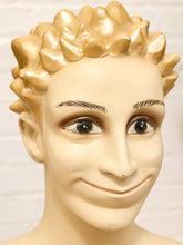 Load image into Gallery viewer, A novelty mannequin head with a glossy beige face, realistic brown eyes, and a wide smile, topped with metallic gold spiky hair, shown against a white brick wall background.
