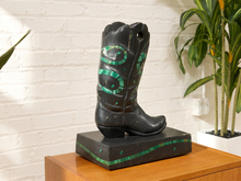 Load image into Gallery viewer, A black decorative cowboy boot sculpture with green inlay details, displayed on a wooden pedestal against a white brick wall with a potted plant in the background.
