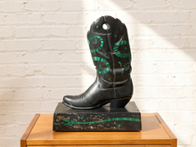 Load image into Gallery viewer, A black cowboy boot sculpture with green painted circular patterns, displayed on a matching black and green pedestal atop a wooden table against a white brick wall.
