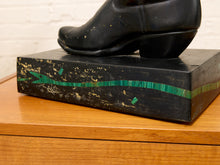 Load image into Gallery viewer, A black cowboy boot sculpture displayed on top of a black rectangular pedestal with green and gold painted stripes, set on a wooden surface against a white brick wall.
