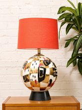 Load image into Gallery viewer, Postmodern ceramic table lamp with rounded abstract painted base in black, pale blue, gold and red, black pedestal, and bright red drum shade
