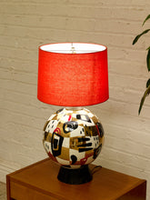 Load image into Gallery viewer, A table lamp with a bright red fabric drum shade and a glossy ceramic base featuring an abstract geometric pattern in gold, black, white, and red, sitting on a wooden side table against a white brick wall.
