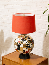 Load image into Gallery viewer, A mid-century modern table lamp featuring a spherical ceramic base with an abstract geometric pattern in beige, black, and red, topped with a solid burnt orange-red drum shade, resting on a wooden side table against a white brick wall.
