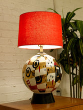 Load image into Gallery viewer, A table lamp with a bright red fabric drum shade and a glossy ceramic base featuring a patchwork of abstract geometric patterns in gold, black, white, and red, sitting on a wooden surface against a white brick wall with a green plant in the background.

