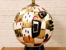 Load image into Gallery viewer, A decorative spherical table lamp with an abstract geometric pattern of gold, white, black, and red panels, sitting on a dark base against a white brick wall.

