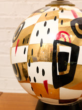 Load image into Gallery viewer, A close-up of a decorative ceramic vase with an abstract geometric pattern featuring gold, white, black, and red shapes, set against a white brick wall background.
